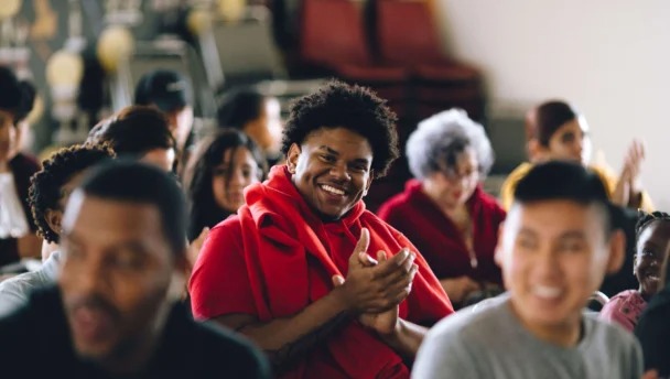 A group of people sit in an indoor setting, smiling and clapping. One person in a red shirt is centered in the image, appearing particularly cheerful.